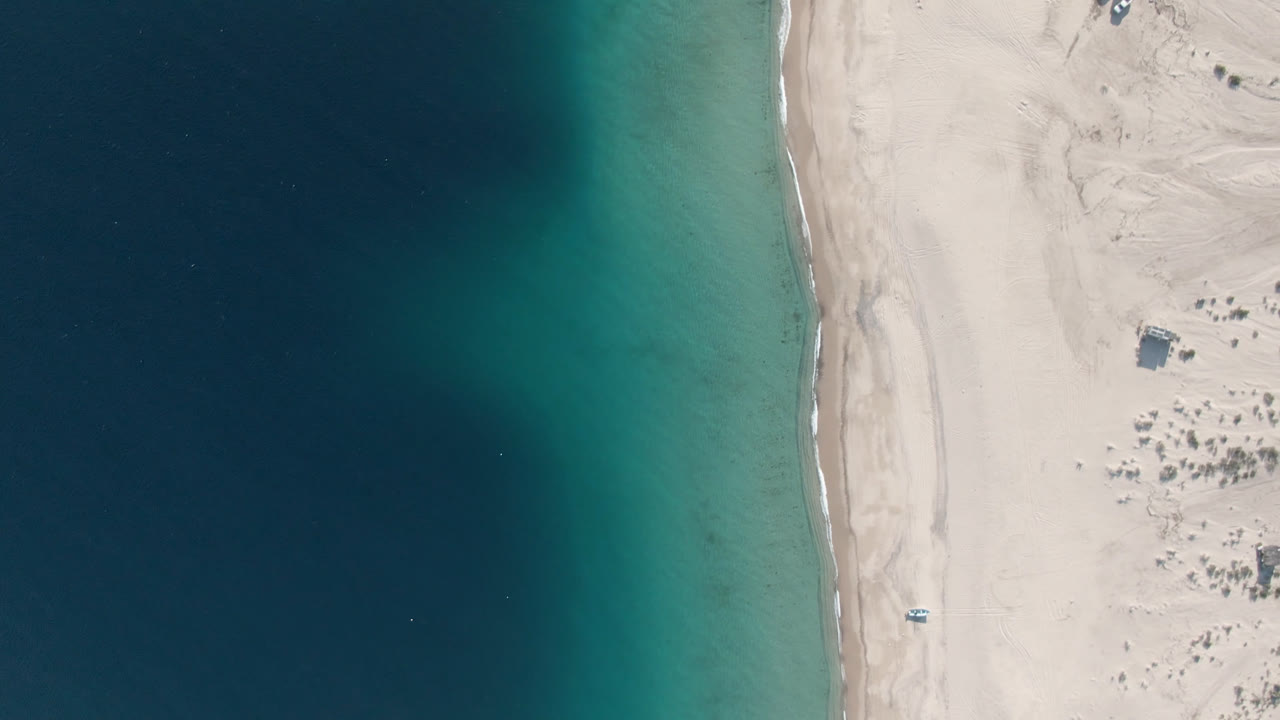 Aerial beach view with turquoise surf edge, pale sand, and scattered small structures inland.