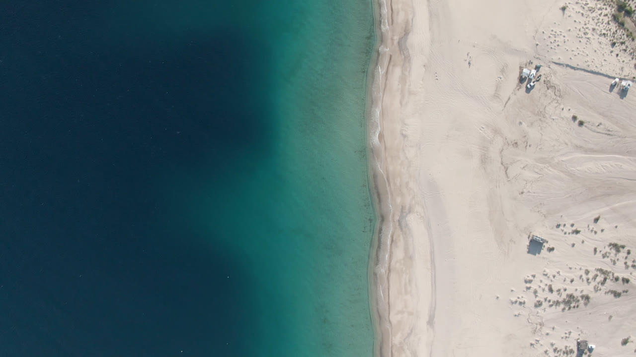 Overhead frame from Los Frailes, Baja California Sur, Mexico showing dark blue water fading into turquoise shallows along a pale sand beach with no visible people.