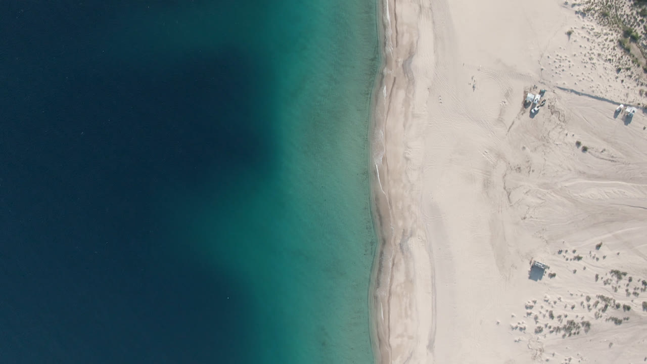 Aerial view over Los Frailes shoreline with turquoise water on the left, pale beach through the center, small objects on the sand, and no visible people.