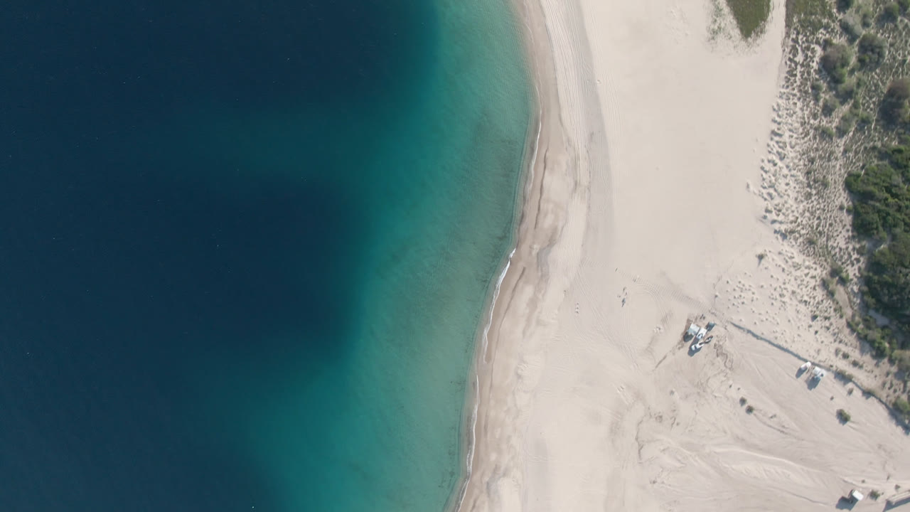 Overhead frame from Los Frailes, Baja California Sur, Mexico showing deep blue water at left, turquoise shallows, pale sand, dune scrub, and small boats near the lower beach with no visible people.