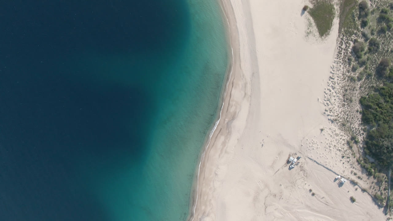 One second into the Los Frailes aerial clip, the frame shows more of the central sandbar-like beach edge between dark blue water and turquoise shallows, with scrub vegetation above and no visible people.