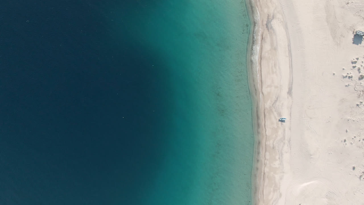 Second overhead frame of Los Frailes shoreline with the beach strip centered between darker offshore water and lighter shallows, with no visible people.
