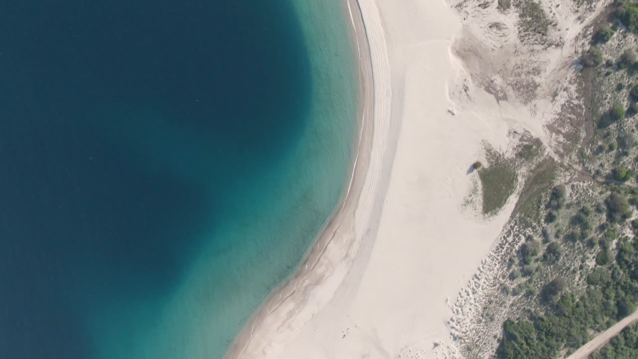 Overhead aerial frame of Los Frailes with dark blue water, a turquoise shallows band, pale curved sand, scrubby dunes, and no visible people.