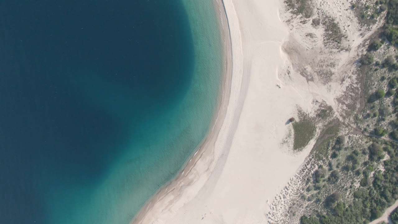 A later overhead frame of Los Frailes shows the beach arc slightly shifted, with green-blue shallows on the left, bright sand in the center, dune scrub on the right, and no visible people.