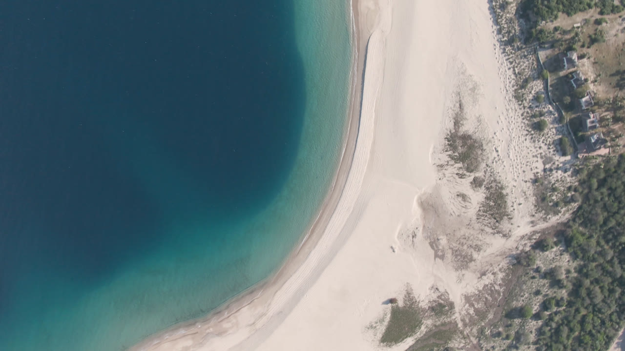 Overhead frame from Los Frailes, Baja California Sur showing a curved pale sand beach, dark blue water, turquoise shallows, trees, small shoreline buildings, and no visible people.