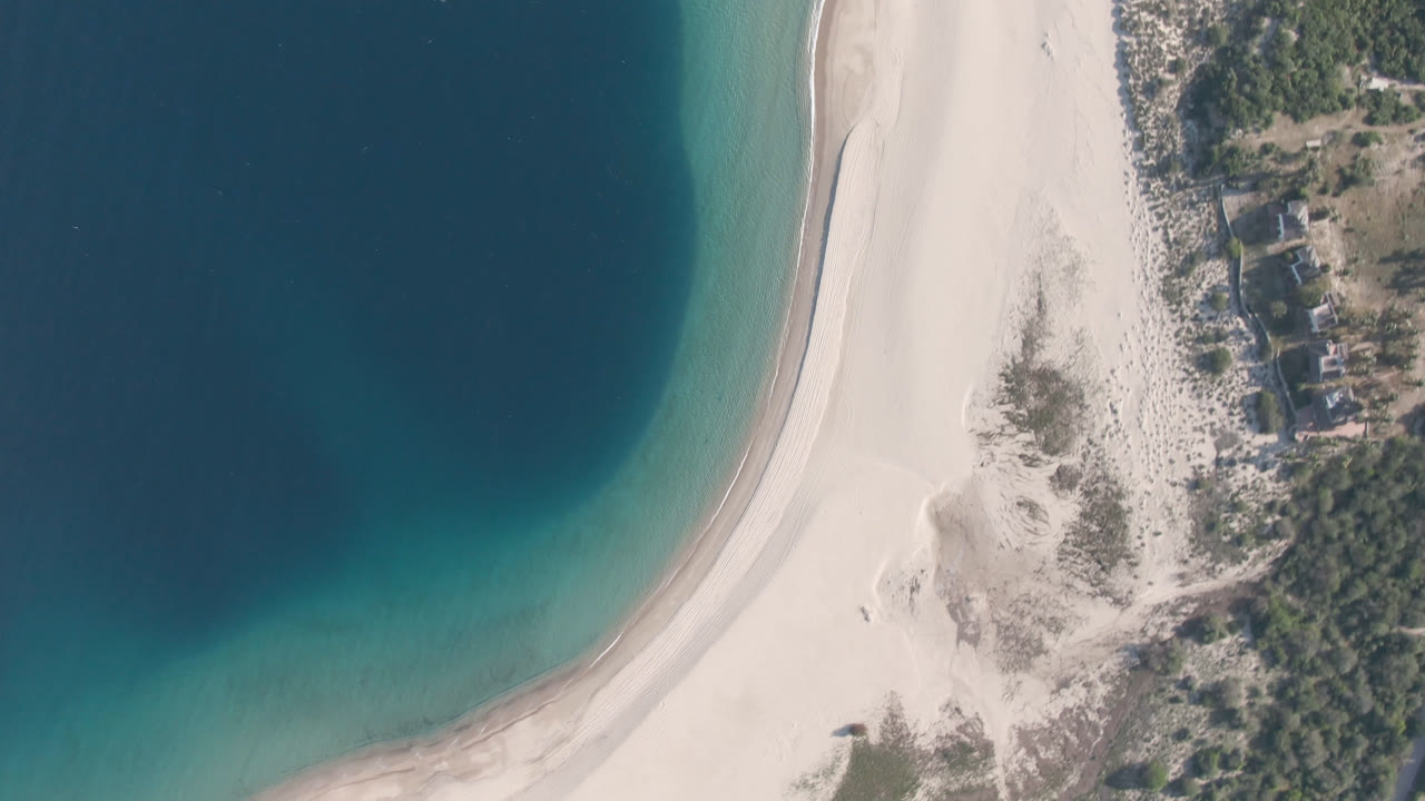 A high aerial view over Los Frailes shows dark water along a pale sand shoreline, a narrow turquoise edge, coastal vegetation, scattered small buildings, and no visible people.