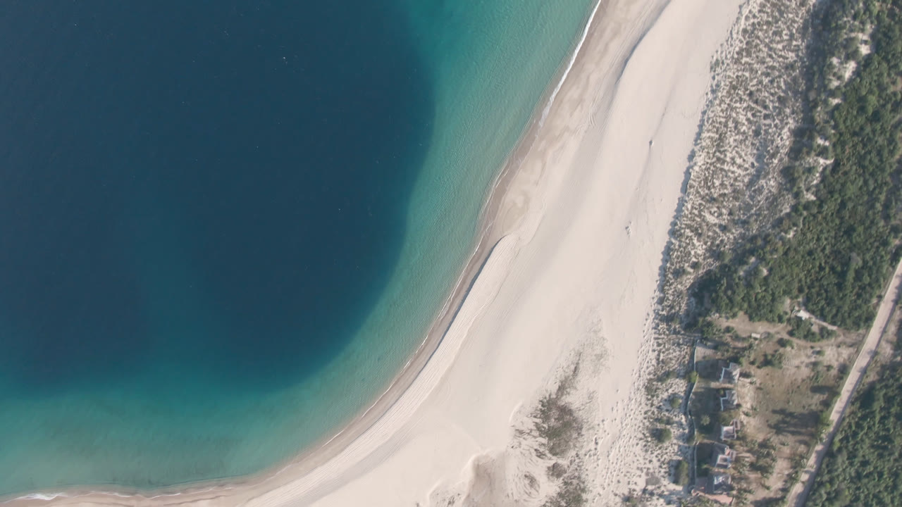 Opening overhead frame of Los Frailes, Baja California Sur, with deep blue water at left, pale sand curving through the center, dune vegetation and small buildings at right, and no visible people.