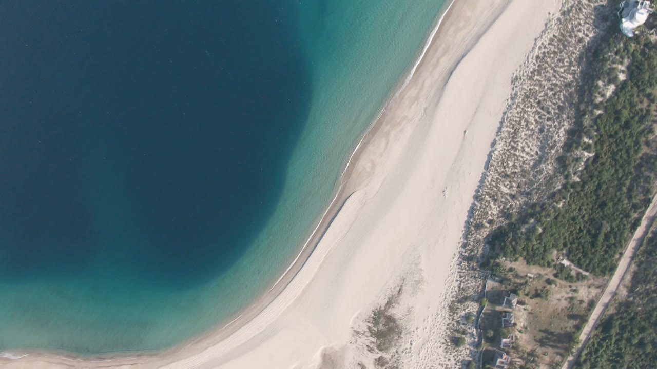 At 0:01, the Los Frailes aerial keeps the water on the left and the sand arc through the middle, with dune scrub and a cluster of small buildings near the lower right, and no visible people.