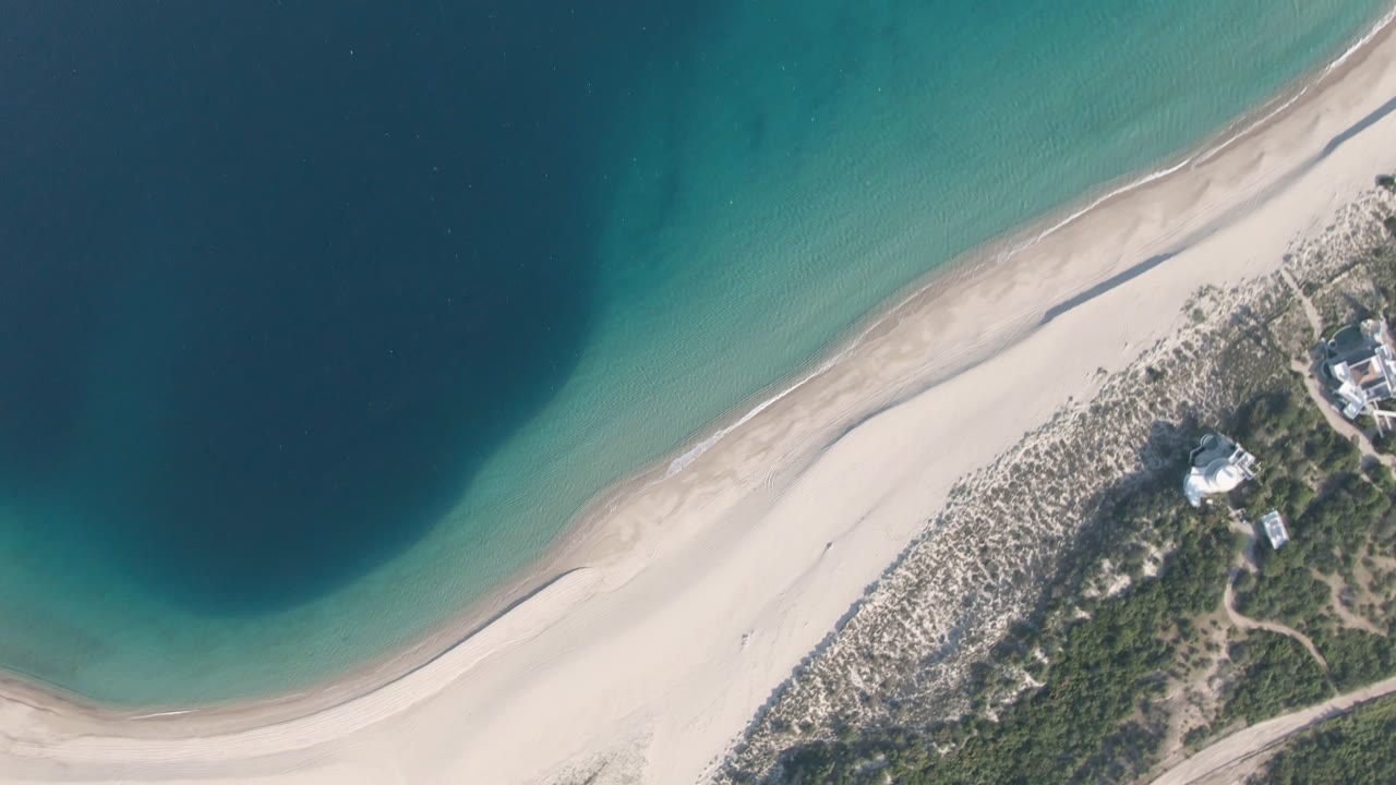 Aerial frame over Los Frailes showing the beach curve shifted slightly across the view, with turquoise water above the sand, scrubland below, and no visible people.