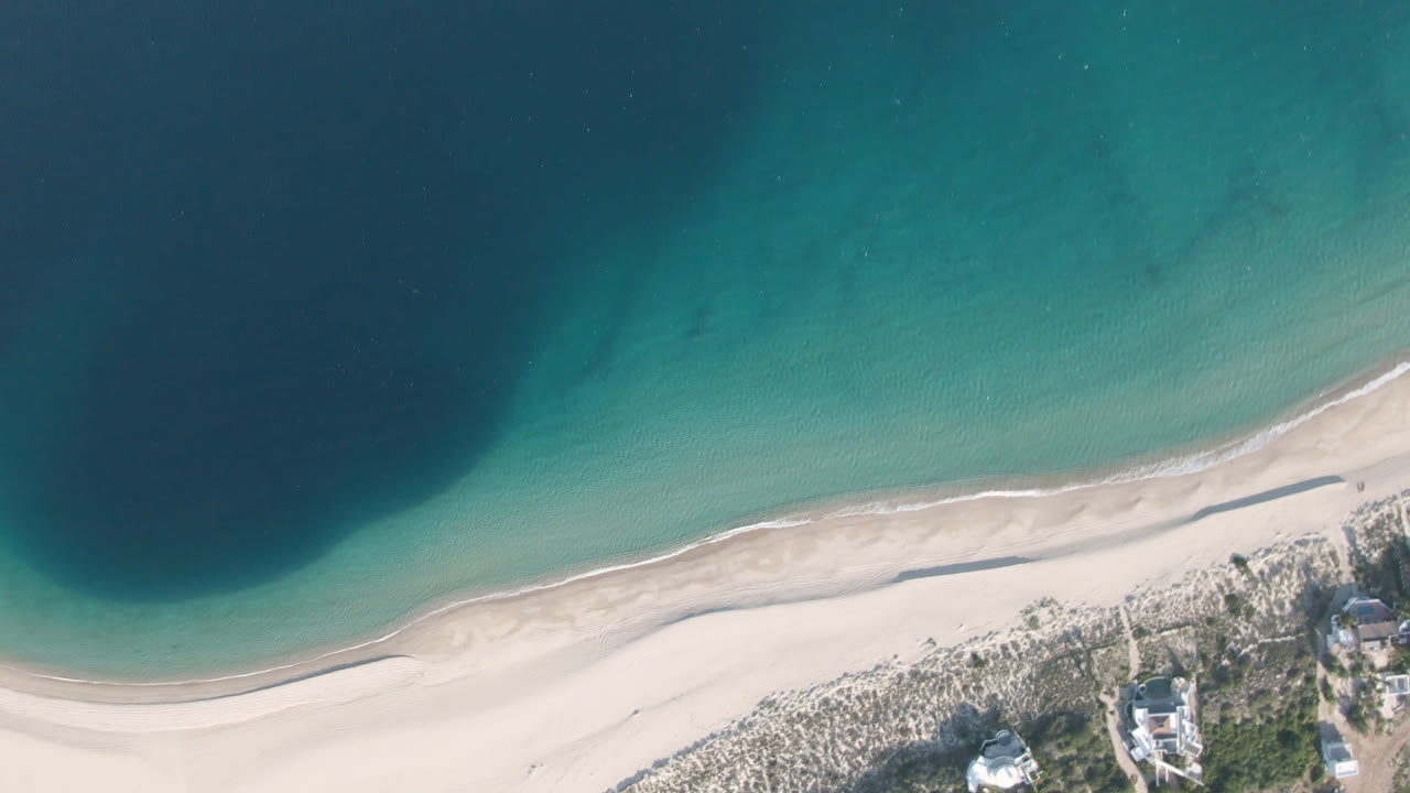 Overhead view in Los Frailes showing the beach curve slightly farther along, with clear turquoise water, a narrow wave edge, dune scrub, small structures, and no visible people.