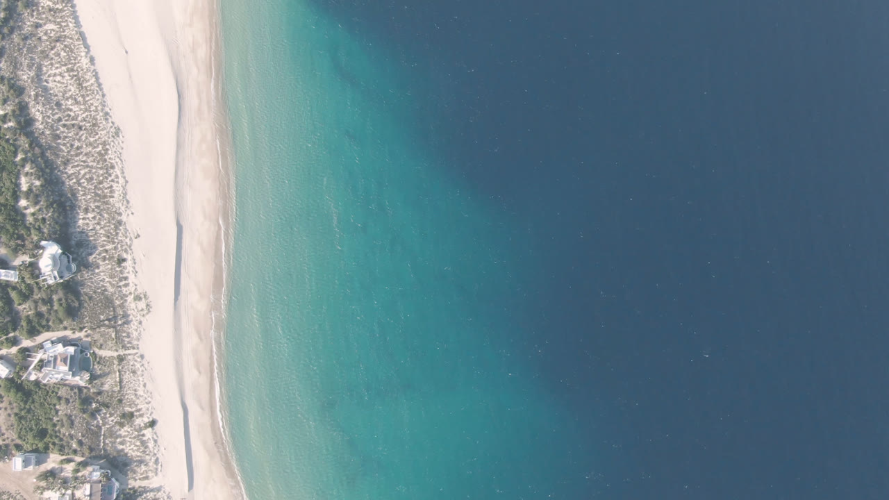 Aerial frame over Los Frailes showing a narrow pale beach, clear turquoise nearshore water, darker offshore water, coastal scrub, and no visible people.
