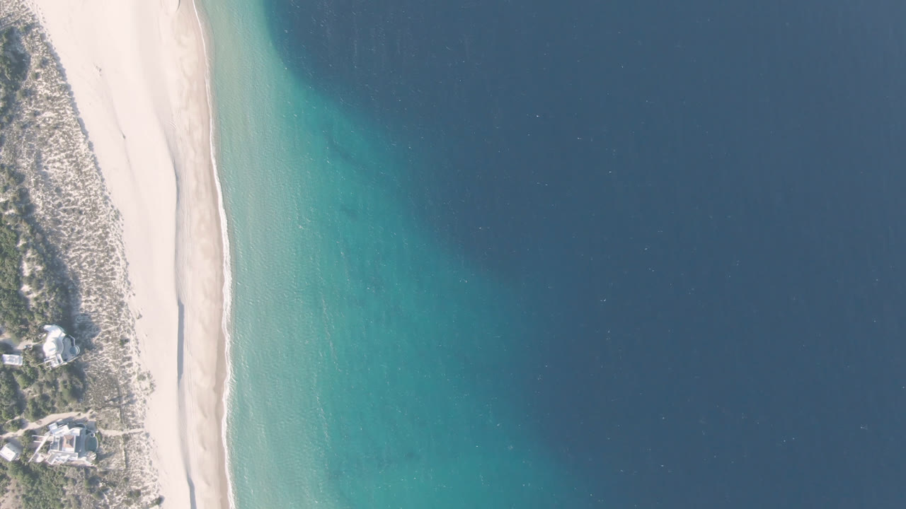 Overhead view of Los Frailes shoreline with pale sand, turquoise shallows, darker blue water, dune vegetation, a few coastal buildings, and no visible people.