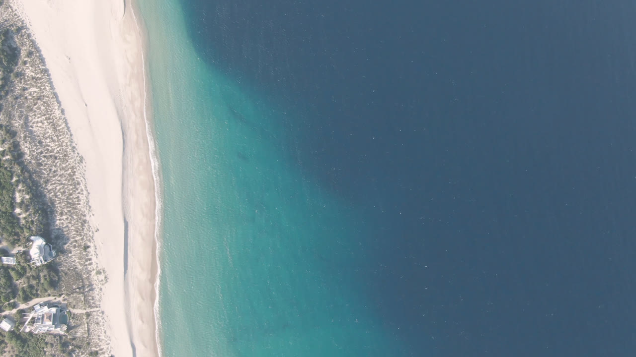 A near-vertical aerial frame over Los Frailes shows the sandy beach centered between clear blue water and scrub-covered dunes with two coastal buildings, with no visible people.