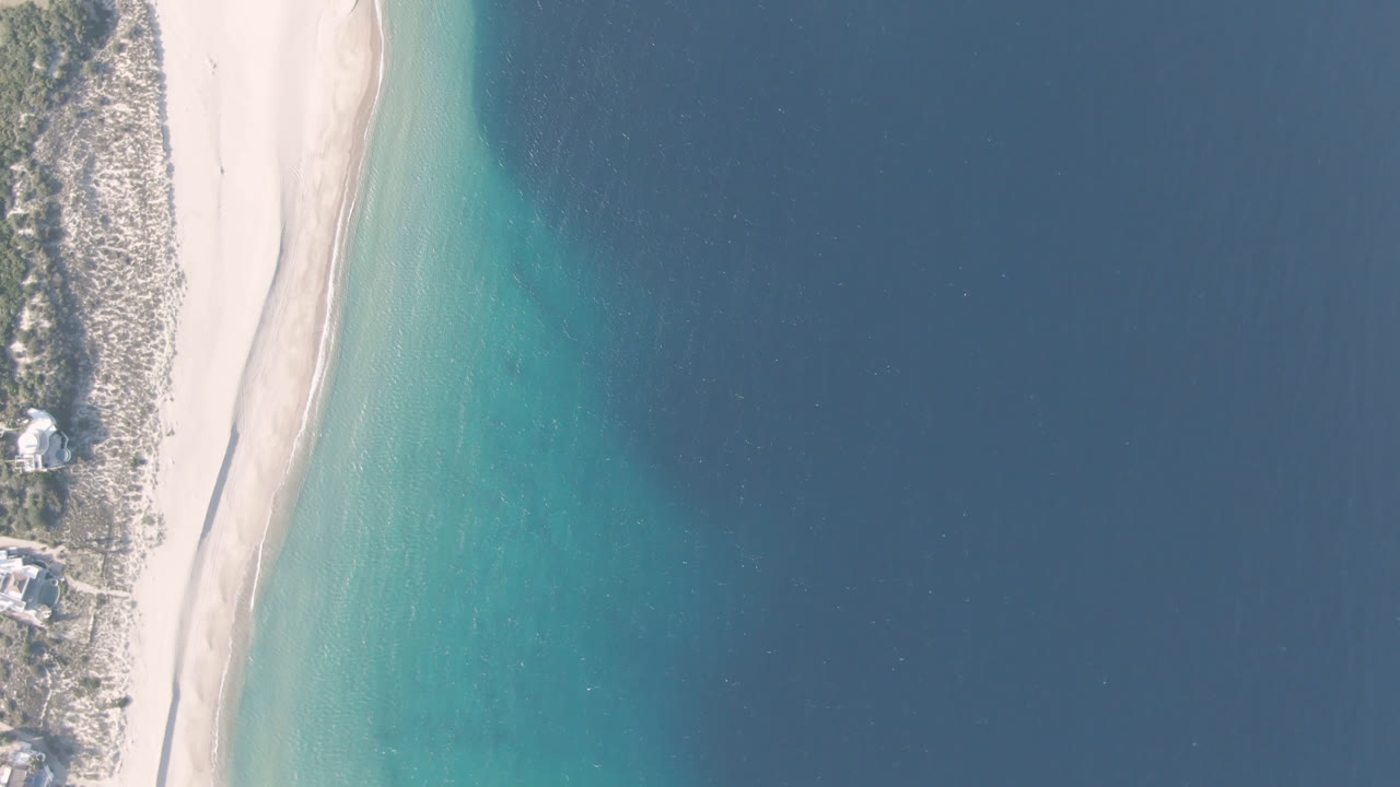 High overhead view of Los Frailes beach in Baja California Sur, Mexico, with pale sand between scrubland and blue water and no visible people.