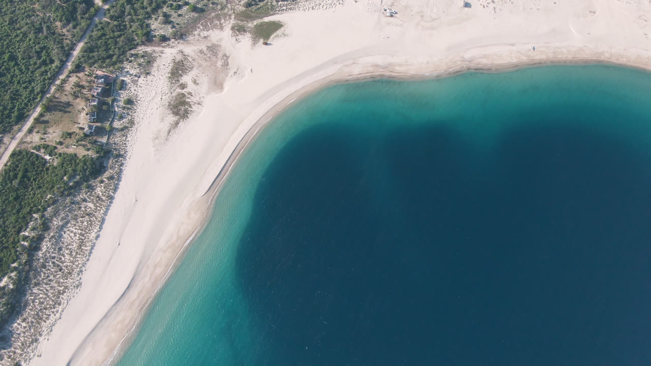 Overhead frame of Los Frailes, Baja California Sur, showing a curved pale sand beach, turquoise shallows, deep blue water, scrub vegetation, a dirt road, small structures, and no visible people.
