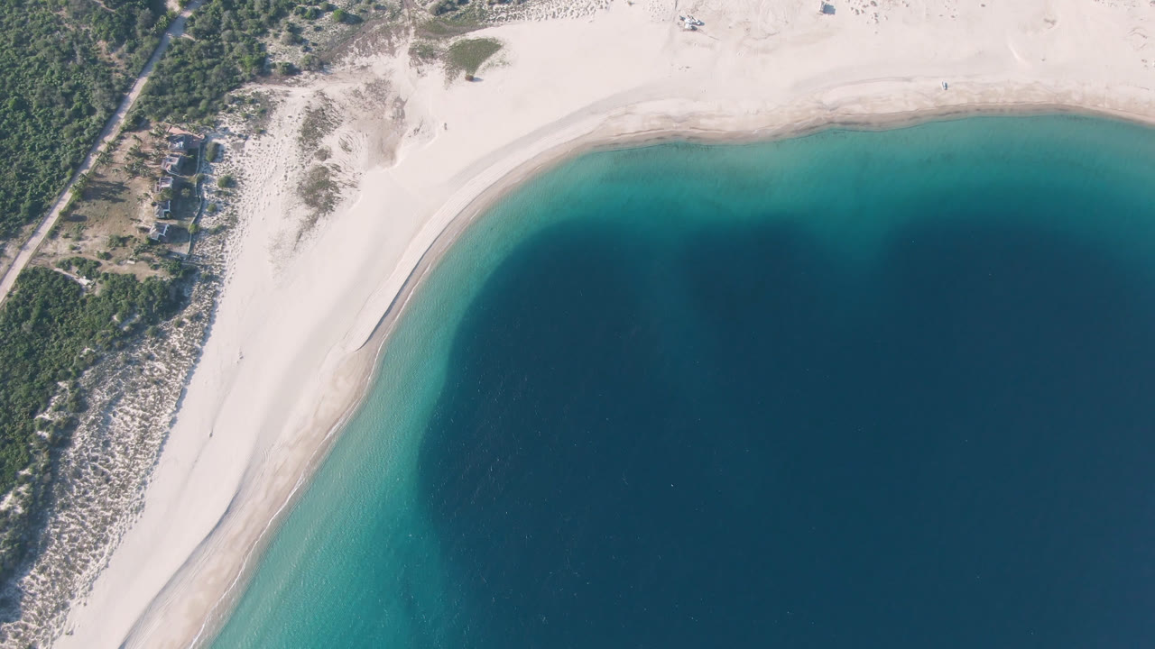 Aerial view over Los Frailes with the sandy shoreline arcing around clear turquoise water, darker bay water beyond, green brush along the edge, and no visible people.