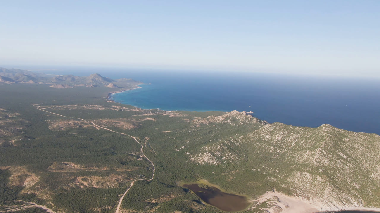 Aerial view over Los Frailes, Baja California Sur, with rocky green hills in the foreground, pale roads across coastal flats, a curved blue bay, open ocean, clear sky, and no visible people.