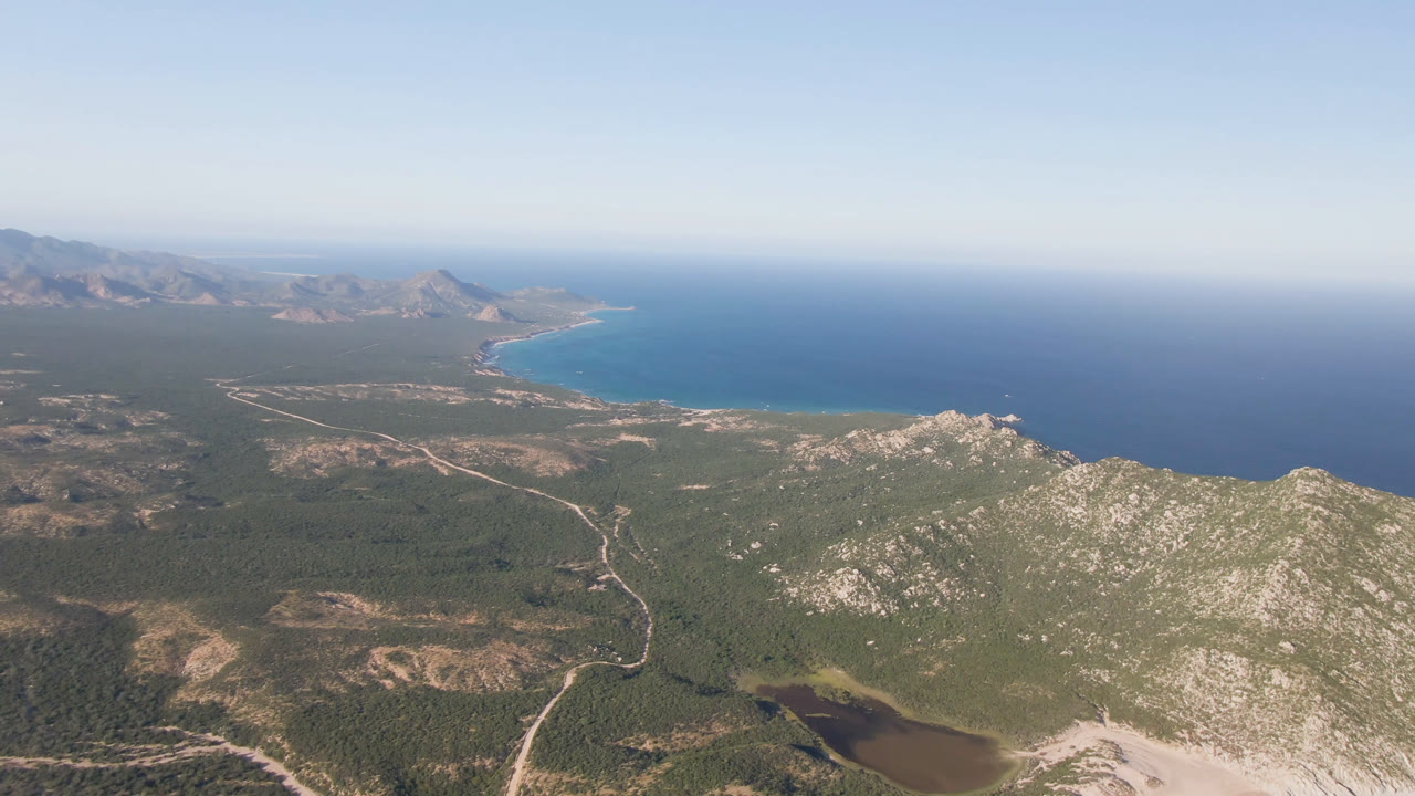 Second aerial frame over Los Frailes with the turquoise nearshore water more centered, rocky ridges across the lower right, coastal flats, dirt roads, distant mountains, and no visible people.