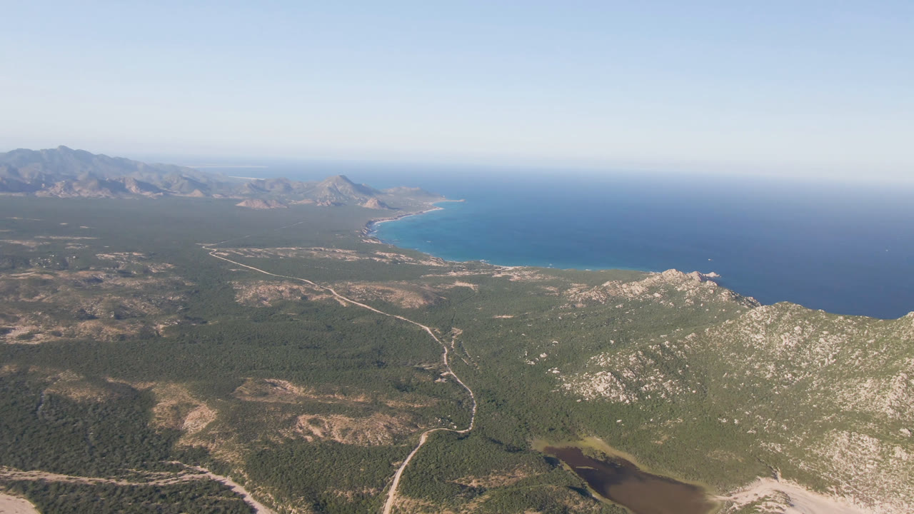 Opening aerial frame over Los Frailes, Baja California Sur, with rocky hills in the foreground, scrubland roads, blue ocean, distant mountains, and no visible people.