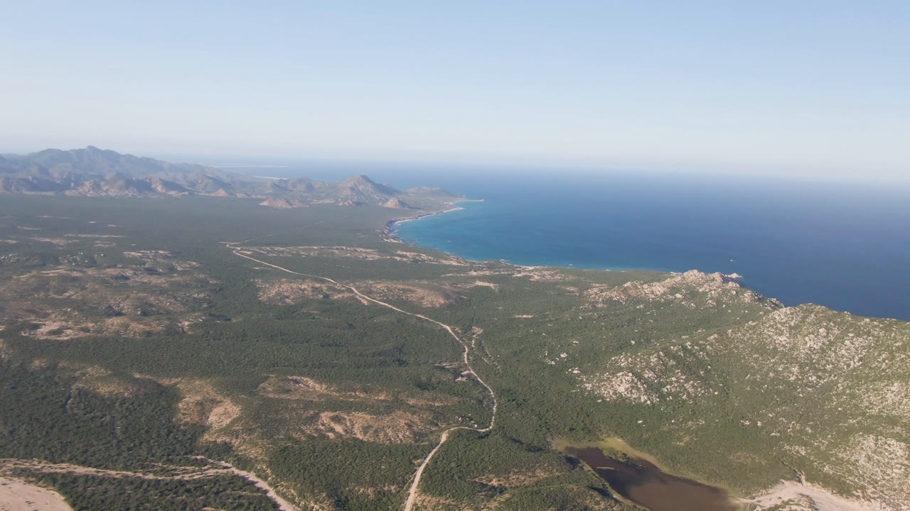 High coastal view with broad water, pale roads, green scrubland, and rocky foreground.