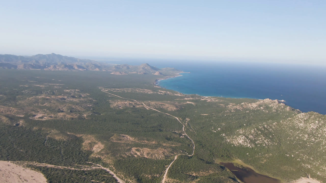 Aerial frame over Los Frailes, Baja California Sur with winding pale roads through green scrubland, rocky hills in front, blue bay at upper right, distant mountains, and no visible people.