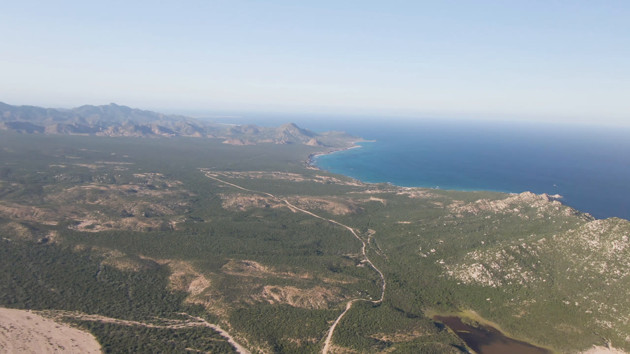 High aerial view in Los Frailes, Baja California Sur showing scrub-covered hills, branching dirt roads, blue coastal water to the right, mountain ridges across the horizon, and no visible people.