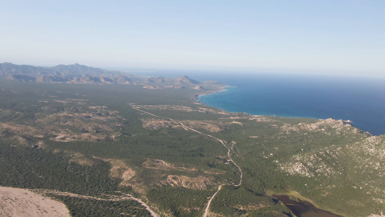 Aerial coastal landscape with rugged hills, road lines, blue sea, and hazy sky.