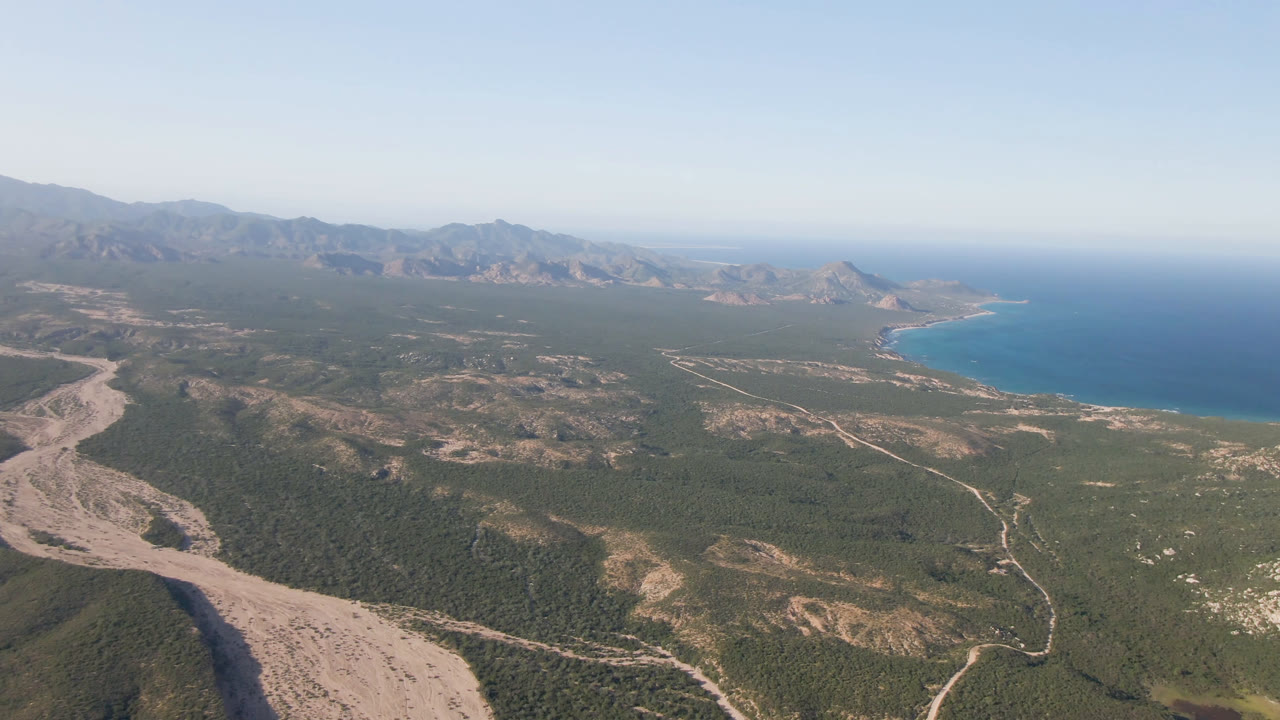 Wide aerial frame from Los Frailes, BCS, Mexico, with a pale arroyo crossing the foreground toward green desert terrain, distant mountains, blue ocean at right, and no visible people.