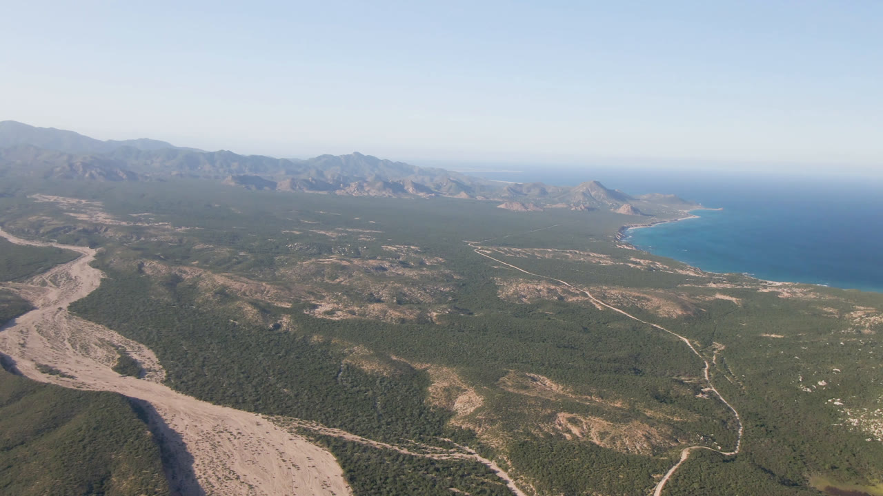 Aerial view over Los Frailes, BCS, Mexico, showing the arroyo nearer the lower left foreground, green scrubland in the center, mountain ridges on the horizon, ocean to the right, and no visible people.