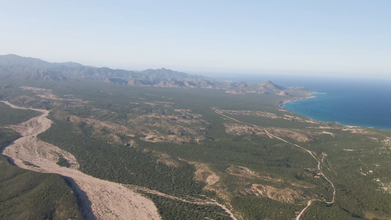 High aerial of desert basin, winding arroyo, mountains, and blue sea