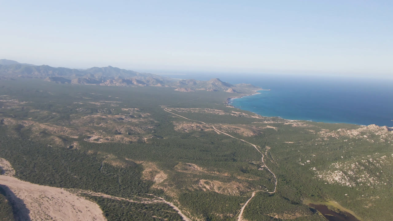Wide aerial frame over San Jose Del Cabo, Mexico, with scrubland and pale dirt roads leading toward a curved blue shoreline and distant mountains, with no visible people.