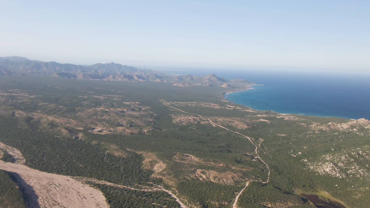 A high aerial view of the San Jose Del Cabo coastline shows dry hills and winding tracks in the foreground, open blue water to the right, and mountains across the horizon, with no visible people.