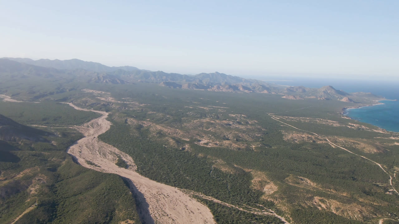Opening aerial frame over Los Frailes, Baja California Sur, with a pale winding road or dry wash crossing green-brown mountain terrain, distant ocean at right, hazy ridges, and no visible people.