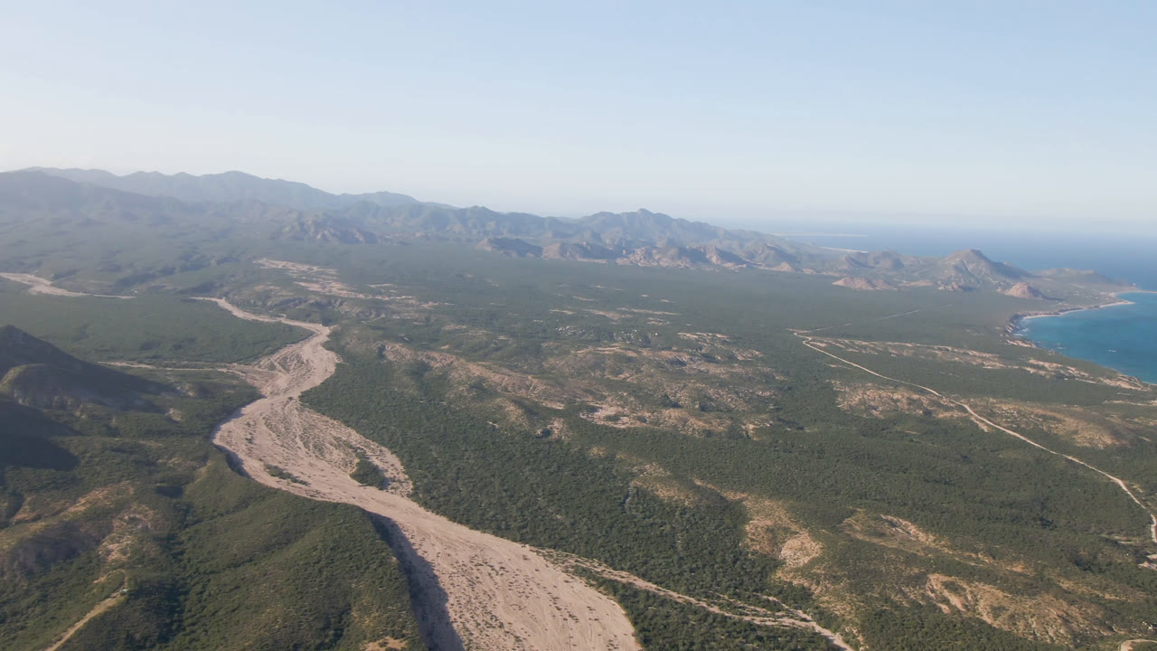 Aerial frame over Los Frailes showing the pale winding corridor lower left, rugged hills across the center, a blue bay on the right edge, hazy mountains in back, and no visible people.