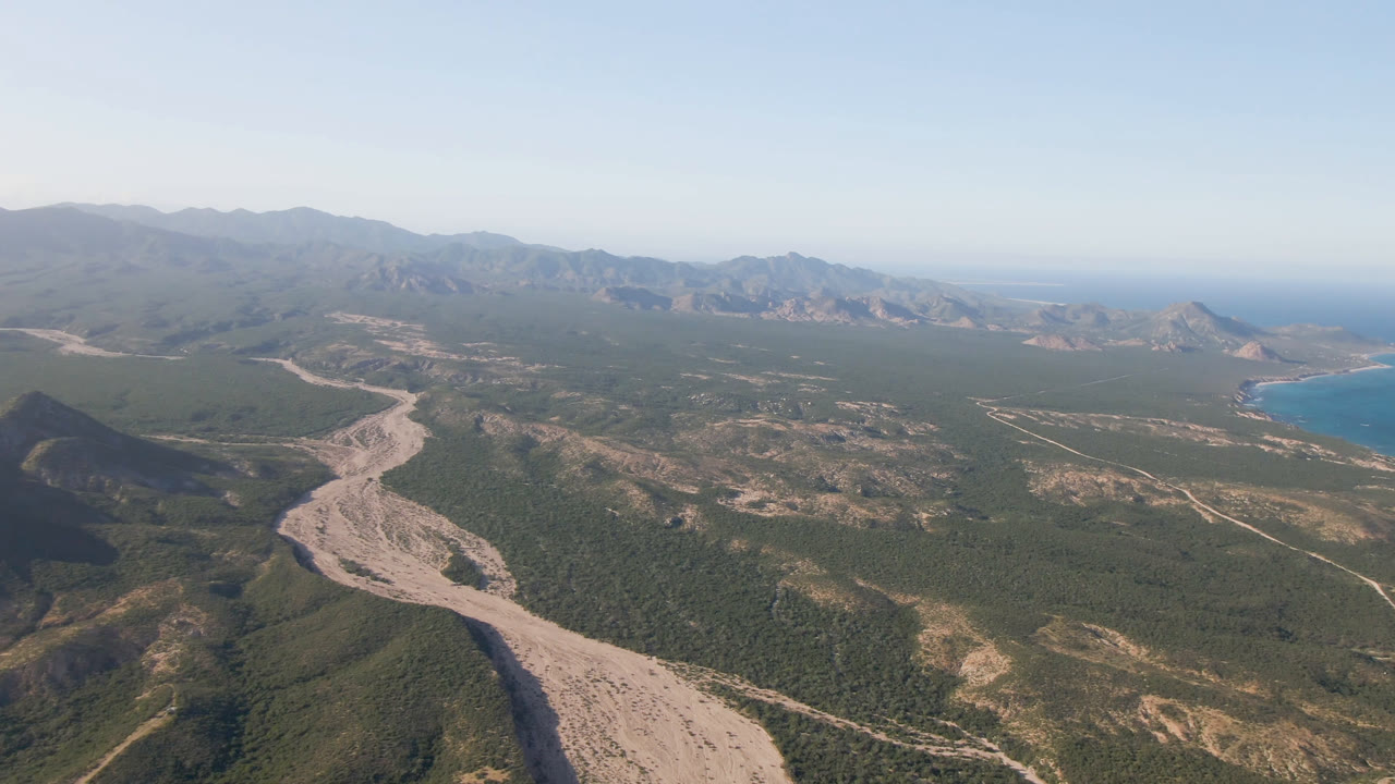 Wide drone view of mountain ridges, a winding pale track, and ocean beyond.