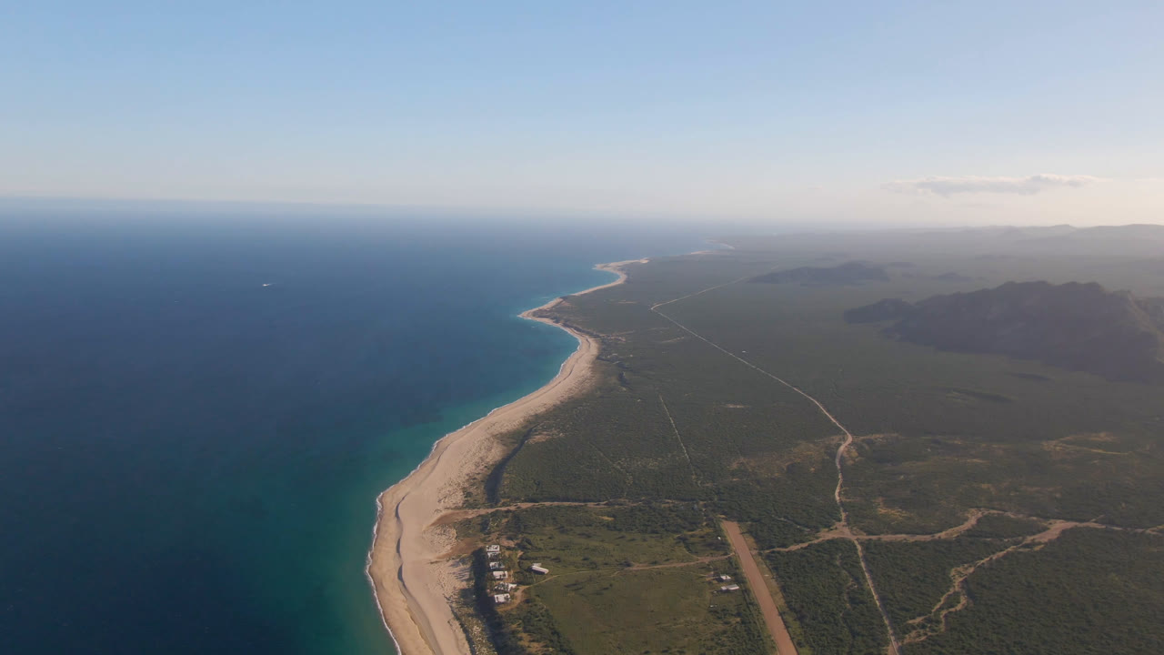 High aerial frame over Los Frailes, Baja California Sur, with deep blue water on the left, a pale beach curving into the distance, inland roads to the right, and no visible people.