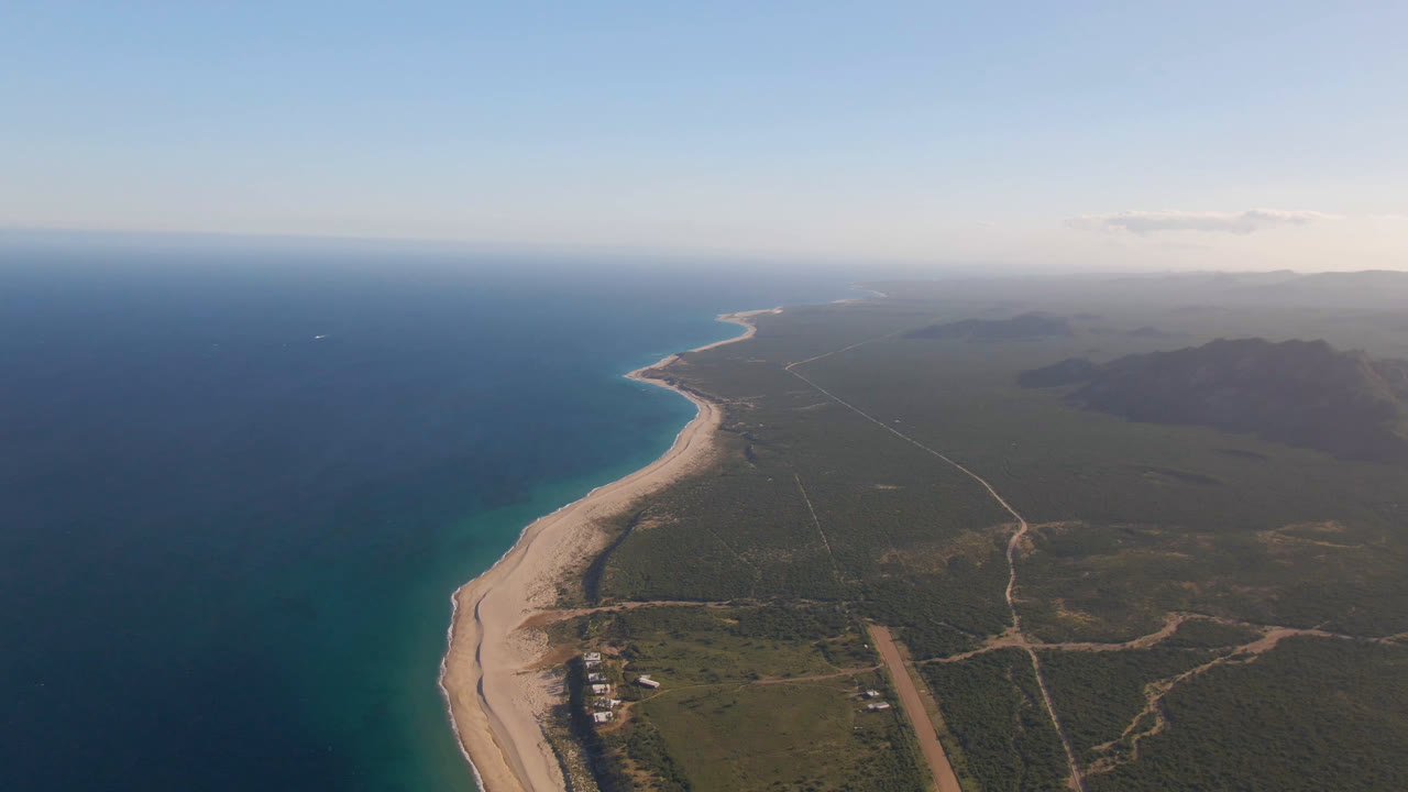 Aerial view at Los Frailes showing the shoreline slightly farther along the curve, with turquoise water near the sand, scrubland roads inland, distant haze, and no visible people.