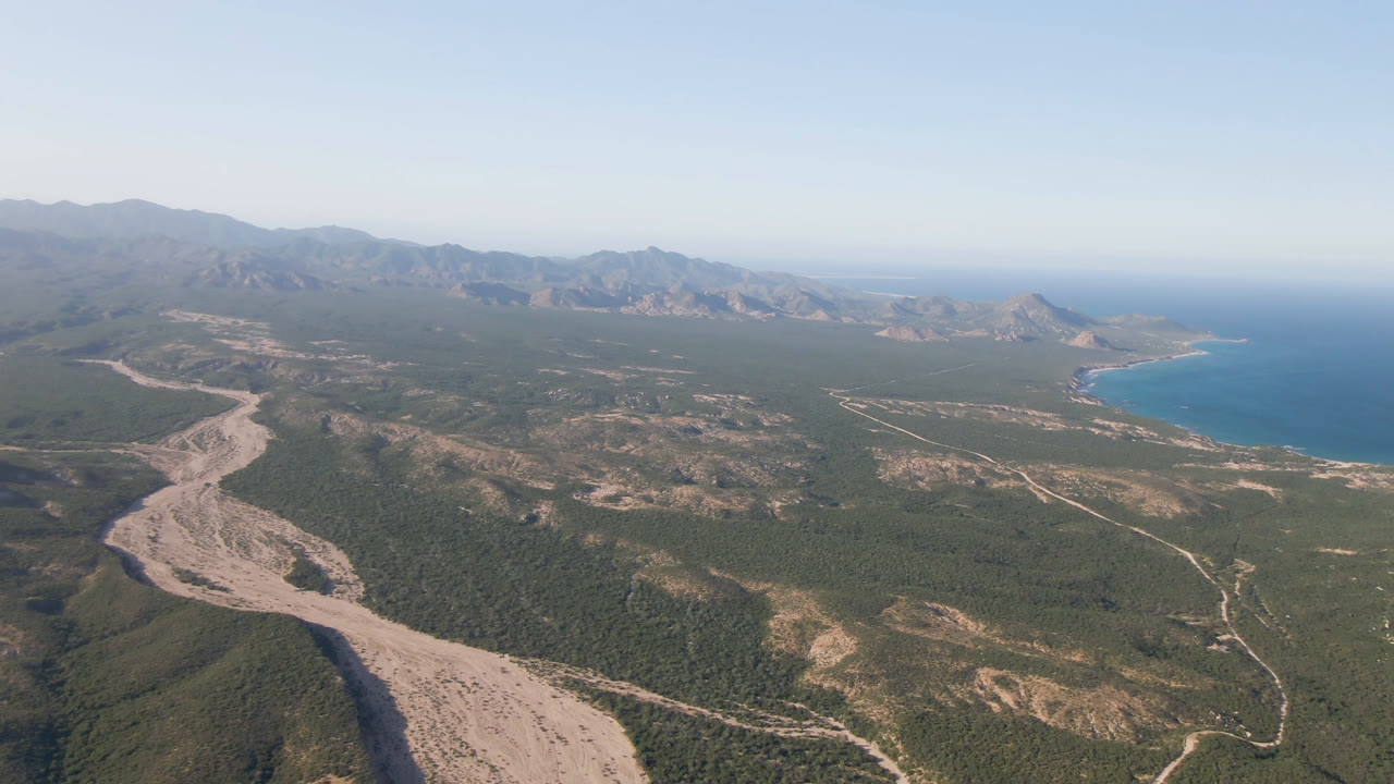 Aerial view over Los Frailes, Baja California Sur, with a pale dry wash in the foreground, scrub hills, winding roads, distant coastline, blue ocean, and no visible people.
