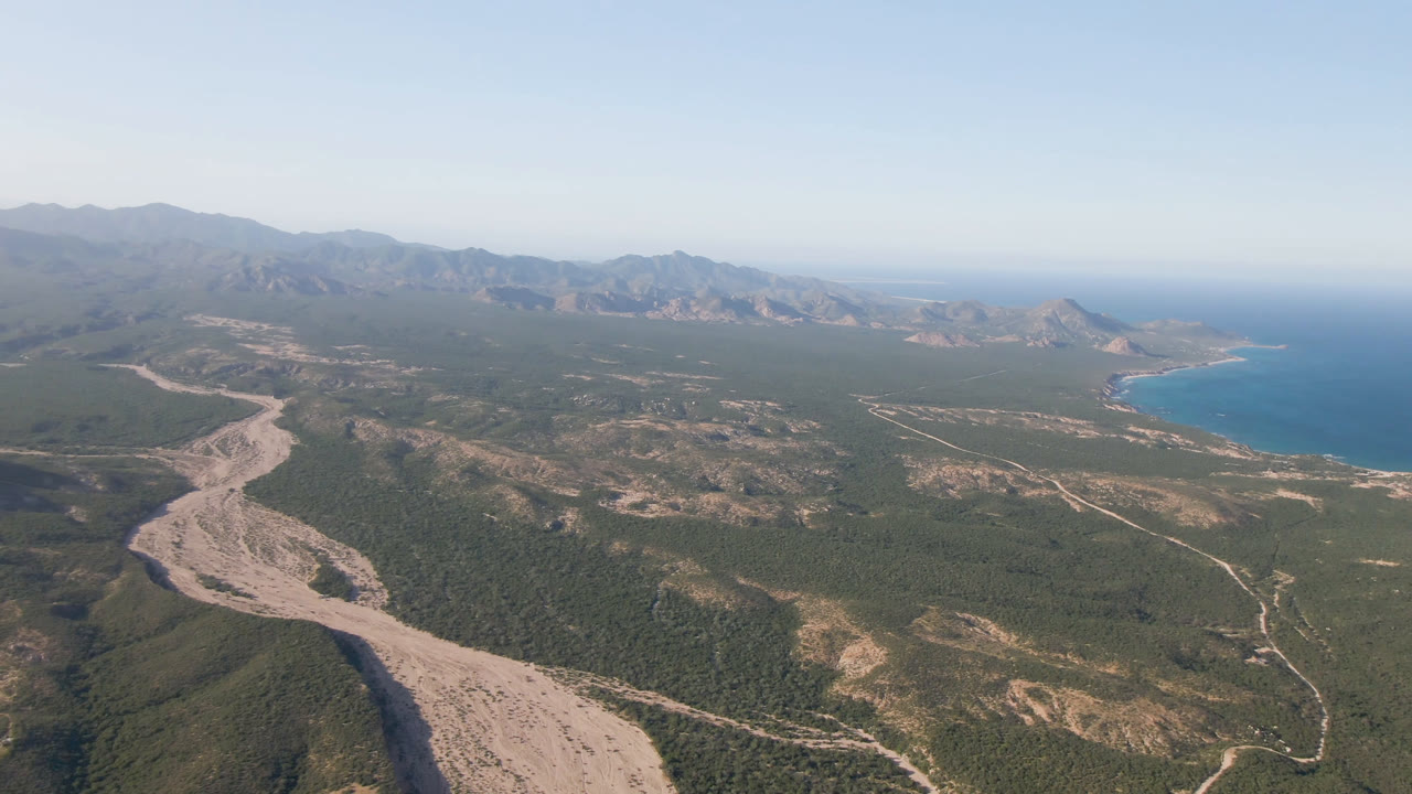Wide aerial frame over Los Frailes showing the dry riverbed more centrally, with green-brown hills, thin roads, distant mountains, blue water, and no visible people.