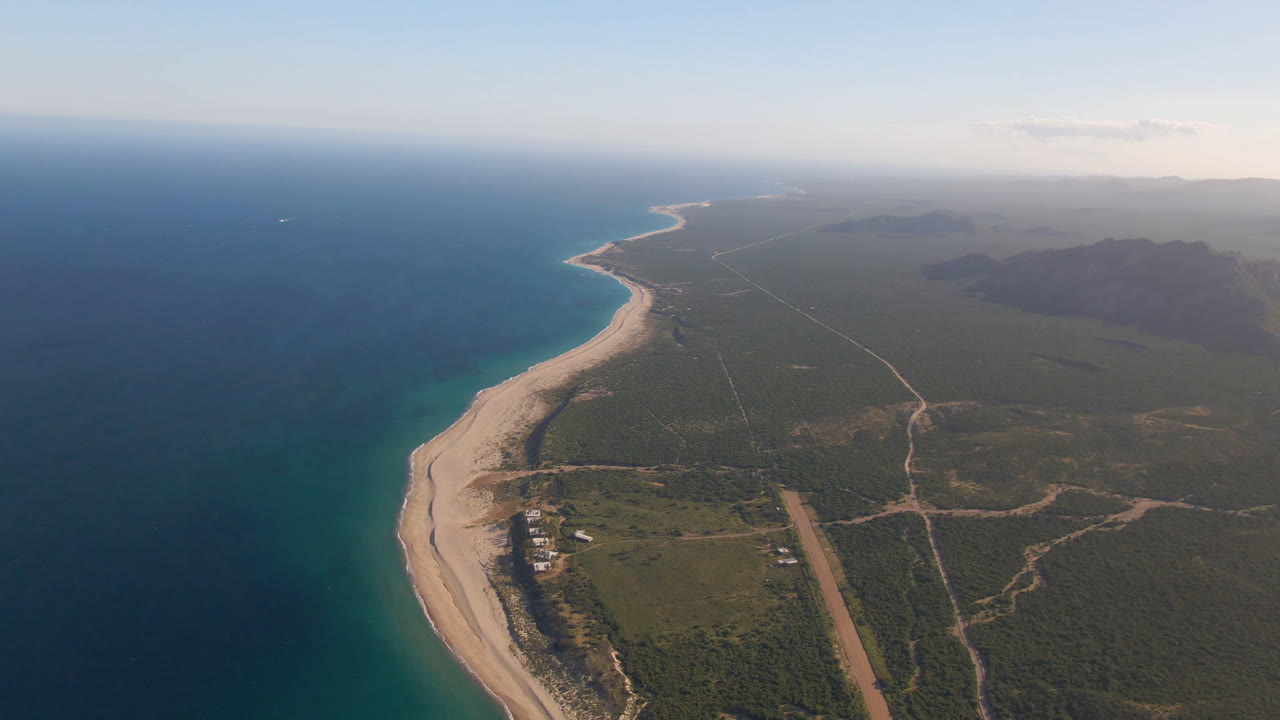 High aerial view over Los Frailes, Baja California Sur, Mexico, with deep blue ocean on the left, a curved sandy beach below, scrubland and dirt roads inland, and no visible people.