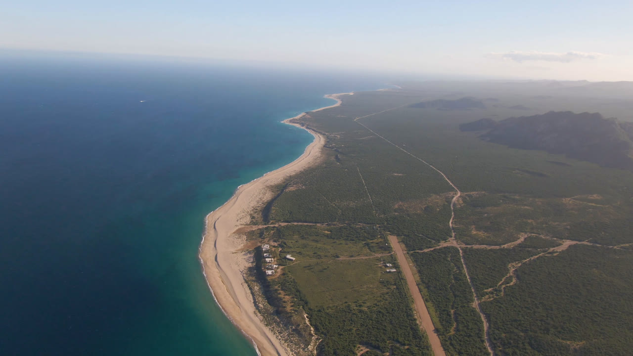 Aerial frame over Los Frailes coastline showing blue water beside a pale sand beach, inland dirt roads and low vegetation, distant hills, hazy sky, and no visible people.
