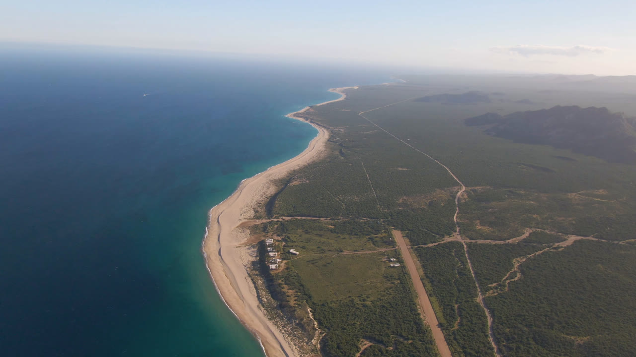 Wide drone-style view of coastline, ocean, beach, roads, scrubland, and distant haze.