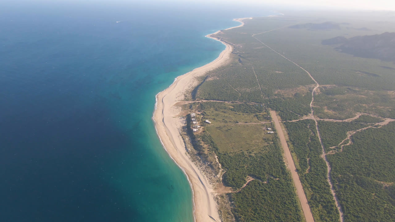 Opening aerial frame over Los Frailes, Baja California Sur, with blue ocean at left, pale beach curving into the distance, inland roads and scrubland to the right, and no visible people.