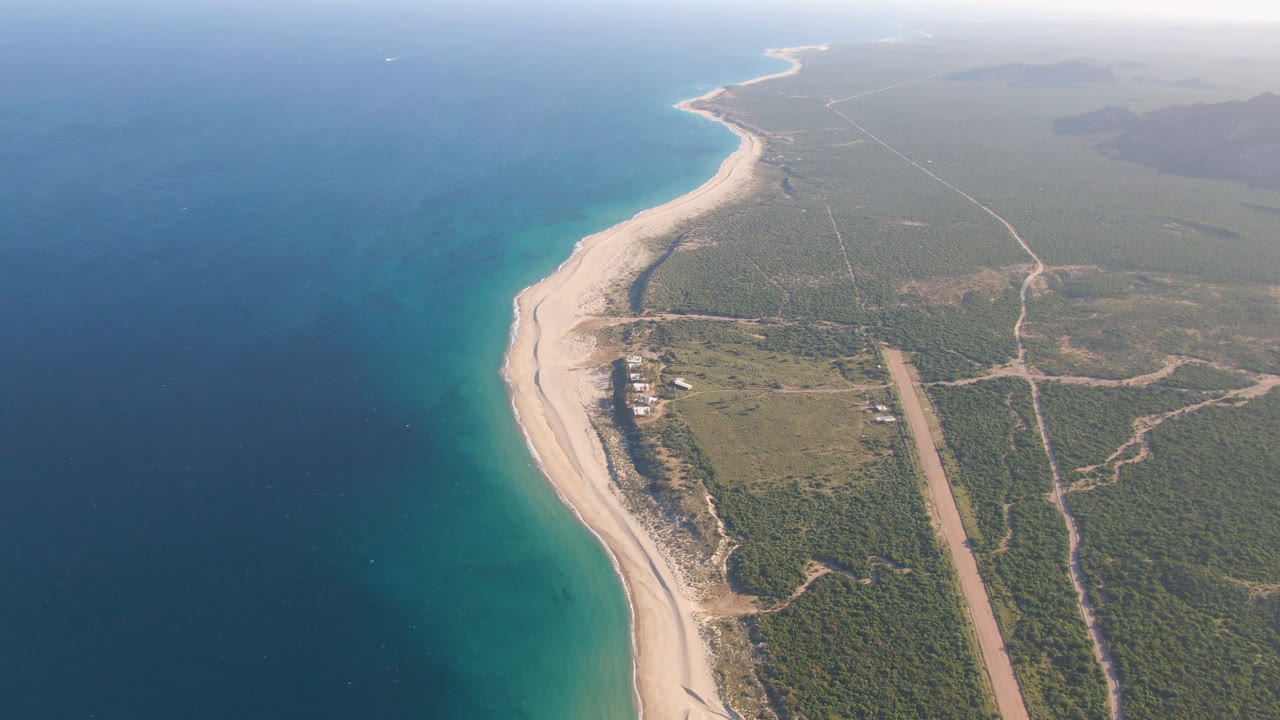 Aerial view of Los Frailes coastline one second in, showing the ocean gradient from dark blue to turquoise, a narrow sand beach, inland vegetation, dirt tracks, and no visible people.