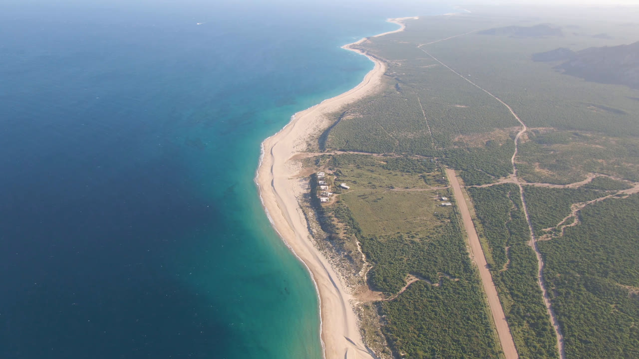 Wide aerial view of beach, ocean, scrubland, and a straight inland road.
