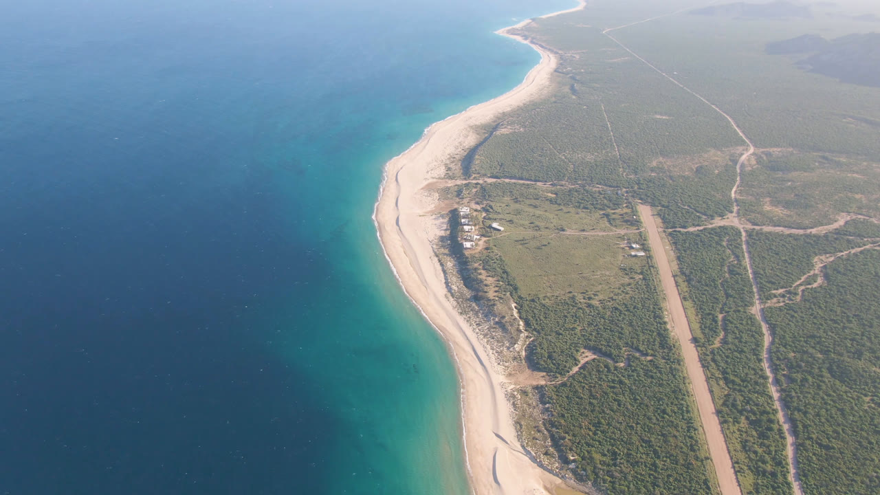 High aerial view over Los Frailes, Baja California Sur, showing a curved pale beach, turquoise shallows, deeper blue water, green scrubland, a straight dirt road, and no visible people.