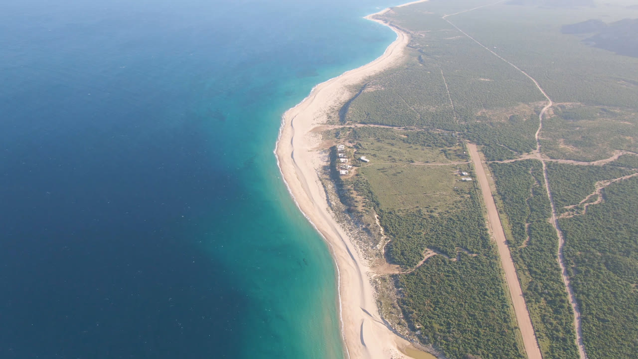 Aerial frame from Los Frailes with the shoreline running diagonally through the image, blue water on the left, inland scrub and dirt tracks on the right, and no visible people.
