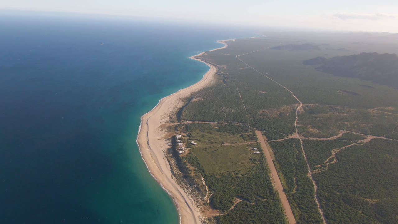Aerial view over Los Frailes, Baja California Sur, Mexico, with deep blue ocean on the left, a curved sandy beach below, inland scrubland and roads to the right, and no visible people.