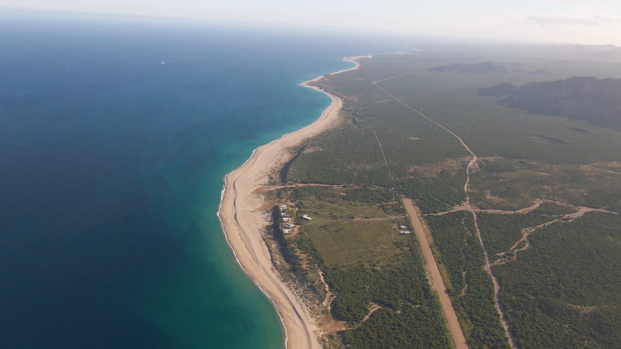 Second aerial frame over Los Frailes shows turquoise shallows along the beach, darker water offshore, a small cluster of buildings near the sand, inland roads, and no visible people.