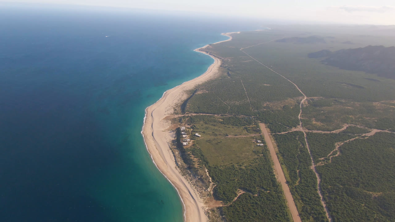 Wide aerial of ocean, beach, coastal vegetation, roads, and distant shoreline.